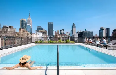 a woman in a hat sitting on the edge of a pool with a city skyline at Blueground Chelsea pool nr The Vessel NYC-2144 in New York